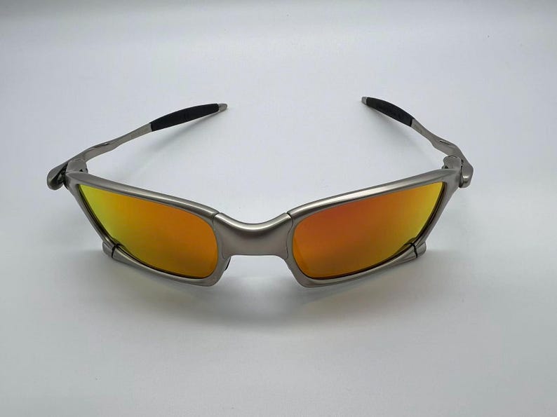 Puede incluir: Gafas de sol con montura plateada y lentes tintadas en naranja. Las gafas tienen un dise&ntilde;o moderno y angular con detalles negros en las patillas. Las lentes son de color naranja vibrante.