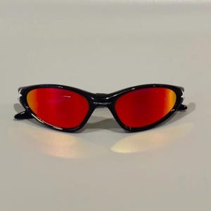 May include: Black sunglasses with red lenses. The sunglasses have a sleek, curved design with a glossy black frame. The lenses have a gradient effect, transitioning from red to orange.