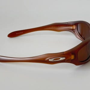 May include: Brown sunglasses with a curved frame and tinted lenses. The sunglasses have a silver logo on the side and are designed for eye protection. The arms of the sunglasses are curved for a comfortable fit.