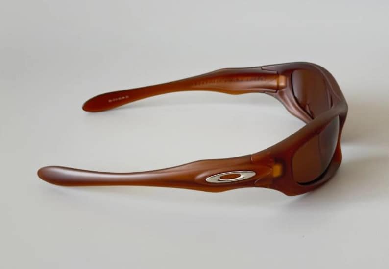 May include: Brown sunglasses with a curved frame and tinted lenses. The sunglasses have a silver logo on the side and are designed for eye protection. The arms of the sunglasses are curved for a comfortable fit.