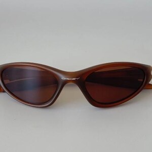 May include: Brown sunglasses with a curved frame and dark lenses. The sunglasses have a sleek, modern design. The brand name "Oakley" is visible on the frame. The sunglasses are set against a plain background.