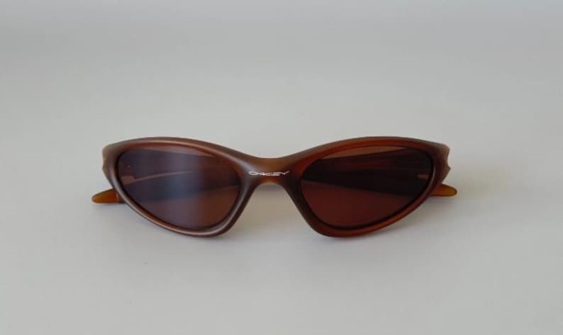 May include: Brown sunglasses with a curved frame and dark lenses. The sunglasses have a sleek, modern design. The brand name "Oakley" is visible on the frame. The sunglasses are set against a plain background.