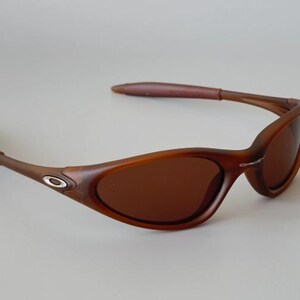 May include: Brown sunglasses with a curved frame and tinted lenses. The sunglasses have a dark brown frame with a silver logo on the side. The arms are a lighter brown color. The lenses are a dark brown color.