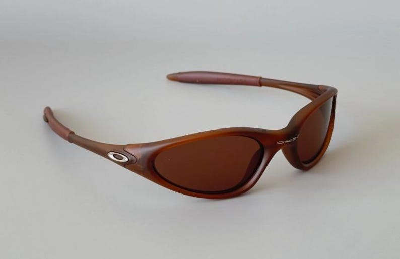 May include: Brown sunglasses with a curved frame and tinted lenses. The sunglasses have a dark brown frame with a silver logo on the side. The arms are a lighter brown color. The lenses are a dark brown color.