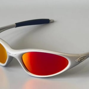 K&ouml;nnte beinhalten: Silberfarbene Sonnenbrille mit rot-orangen Farbverlaufsgl&auml;sern. Die Sonnenbrille hat ein schlankes Design mit schwarzen B&uuml;gelenden. Die Gl&auml;ser sind gew&ouml;lbt und bieten einen lebendigen Kontrast zum silbernen Rahmen. Ein stilvolles Accessoire zum Schutz der Augen.