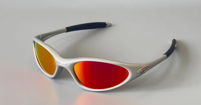 K&ouml;nnte beinhalten: Silberfarbene Sonnenbrille mit rot-orangen Farbverlaufsgl&auml;sern. Die Sonnenbrille hat ein schlankes Design mit schwarzen B&uuml;gelenden. Die Gl&auml;ser sind gew&ouml;lbt und bieten einen lebendigen Kontrast zum silbernen Rahmen. Ein stilvolles Accessoire zum Schutz der Augen.