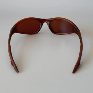 May include: Brown sunglasses with a curved frame and tinted lenses. The sunglasses have a sleek, modern design, with a dark brown color throughout the frame and lenses. The arms of the sunglasses curve down and away from the lenses.