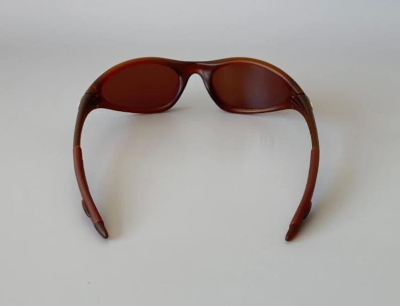 May include: Brown sunglasses with a curved frame and tinted lenses. The sunglasses have a sleek, modern design, with a dark brown color throughout the frame and lenses. The arms of the sunglasses curve down and away from the lenses.