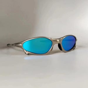 May include: Silver-framed sunglasses with blue mirrored lenses. The sunglasses have a sleek, modern design with a curved frame and a subtle nose bridge. The lenses reflect a vibrant blue hue, offering a stylish and functional accessory.