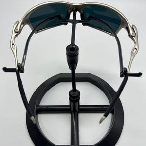 Puede incluir: Gafas de sol con montura plateada y lentes oscuras expuestas en un soporte negro. Las gafas tienen un dise&ntilde;o curvo y un acabado met&aacute;lico. El soporte es circular con un soporte central.
