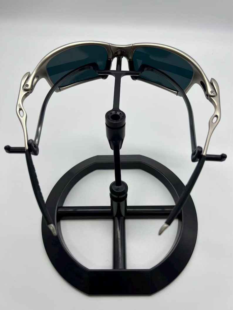 Puede incluir: Gafas de sol con montura plateada y lentes oscuras expuestas en un soporte negro. Las gafas tienen un dise&ntilde;o curvo y un acabado met&aacute;lico. El soporte es circular con un soporte central.
