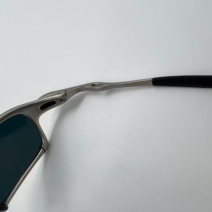 Puede incluir: Primer plano de unas gafas de sol con montura plateada y lentes oscuras. Las patillas son negras y se curvan alrededor de las orejas. Las gafas de sol est&aacute;n sobre un fondo blanco.
