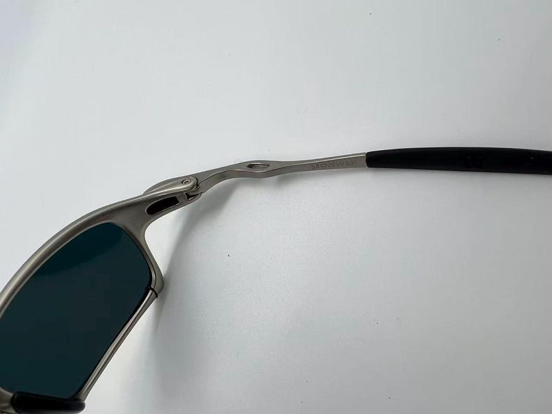Puede incluir: Primer plano de unas gafas de sol con montura plateada y lentes oscuras. Las patillas son negras y se curvan alrededor de las orejas. Las gafas de sol est&aacute;n sobre un fondo blanco.