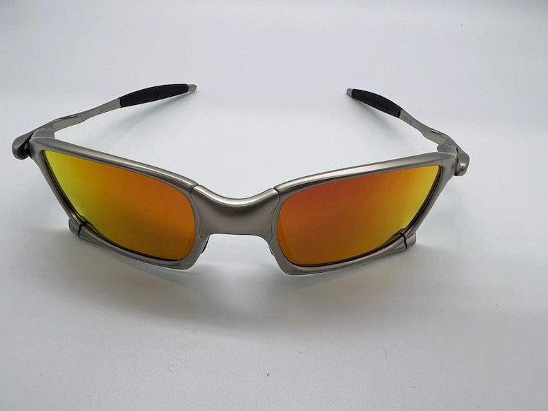Puede incluir: Gafas de sol con montura plateada y lentes naranjas y amarillas. Las gafas de sol tienen un dise&ntilde;o elegante y moderno con una montura curvada y puntas de patillas negras. Las lentes ofrecen un degradado de color.