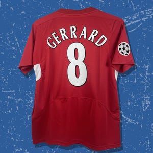 May include: Red soccer jersey with the name "GERRARD" and the number "8" in white. The jersey has white accents on the sleeves and a small emblem on the right sleeve. The jersey is displayed against a blue background.