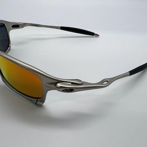 Puede incluir: Gafas de sol plateadas con lentes de color naranja-amarillo y puntas de patillas negras. Las gafas tienen un dise&ntilde;o elegante y moderno, adecuado para actividades al aire libre. Las lentes son reflectantes y la montura es met&aacute;lica.