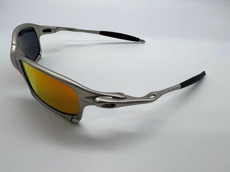 Puede incluir: Gafas de sol plateadas con lentes de color naranja-amarillo y puntas de patillas negras. Las gafas tienen un dise&ntilde;o elegante y moderno, adecuado para actividades al aire libre. Las lentes son reflectantes y la montura es met&aacute;lica.