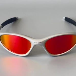 K&ouml;nnte beinhalten: Sonnenbrille mit silbernem Rahmen und leuchtend rot-orangen Gl&auml;sern. Die Brille hat ein schlankes, modernes Design mit schwarzen B&uuml;gelenden. Das Oakley-Logo ist auf dem Rahmen sichtbar. Diese Sonnenbrille eignet sich f&uuml;r Outdoor-Aktivit&auml;ten.