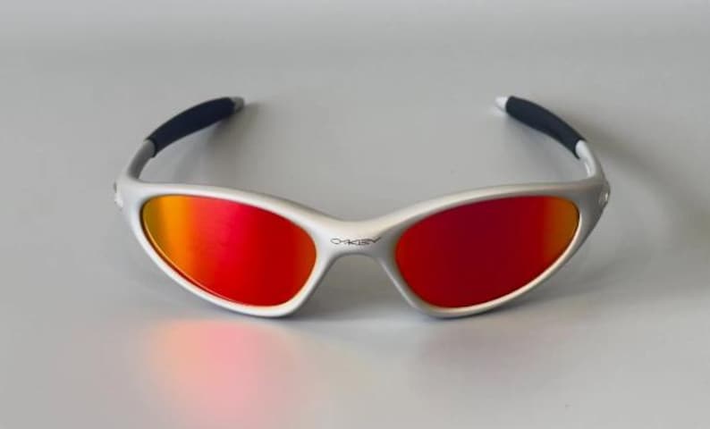 K&ouml;nnte beinhalten: Sonnenbrille mit silbernem Rahmen und leuchtend rot-orangen Gl&auml;sern. Die Brille hat ein schlankes, modernes Design mit schwarzen B&uuml;gelenden. Das Oakley-Logo ist auf dem Rahmen sichtbar. Diese Sonnenbrille eignet sich f&uuml;r Outdoor-Aktivit&auml;ten.