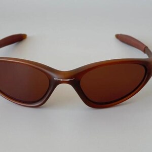 May include: Brown sunglasses with a curved frame and tinted lenses. The sunglasses have a sleek, modern design, with the brand name "Oakley" visible on the frame. The arms are a darker shade of brown.