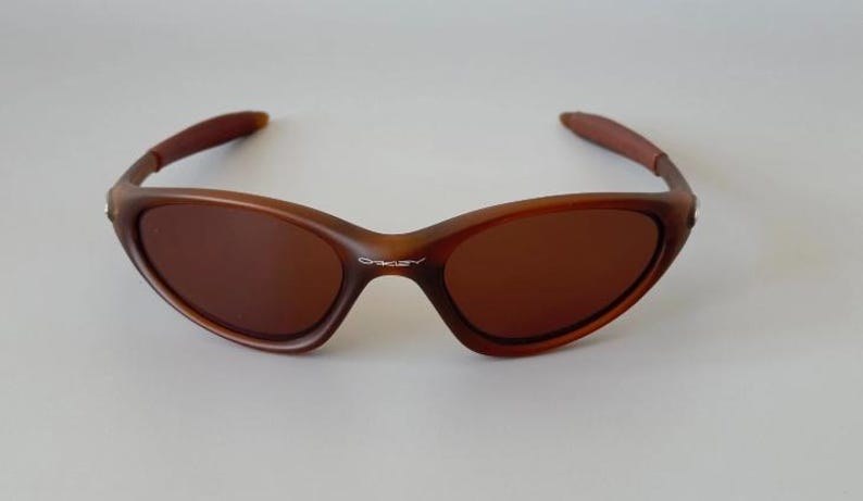 May include: Brown sunglasses with a curved frame and tinted lenses. The sunglasses have a sleek, modern design, with the brand name "Oakley" visible on the frame. The arms are a darker shade of brown.