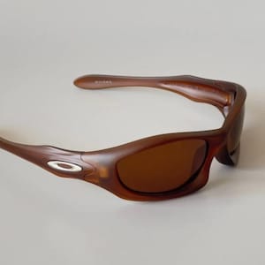 May include: Brown sunglasses with a curved frame and tinted lenses. The sunglasses have a small, oval silver accent on the temples. The lenses are a dark brown color, and the frame is a lighter brown.