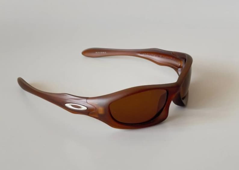 May include: Brown sunglasses with a curved frame and tinted lenses. The sunglasses have a small, oval silver accent on the temples. The lenses are a dark brown color, and the frame is a lighter brown.