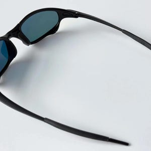 May include: Black sunglasses with blue-tinted lenses. The sunglasses have a sleek, modern design with a curved frame and temples. The lenses are reflective, and the sunglasses are set against a white background.