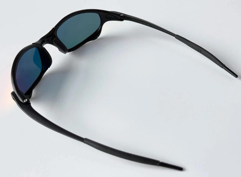 May include: Black sunglasses with blue-tinted lenses. The sunglasses have a sleek, modern design with a curved frame and temples. The lenses are reflective, and the sunglasses are set against a white background.