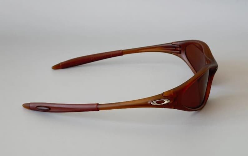 May include: Brown sunglasses with a curved frame and tinted lenses. The temples are a darker brown than the frame and feature a silver logo. The sunglasses are designed for outdoor use.