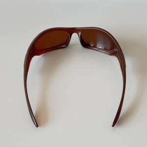 May include: Brown sunglasses with a curved frame and tinted lenses. The sunglasses have a sleek, modern design, with a dark brown color. The arms of the sunglasses curve down and away from the lenses.