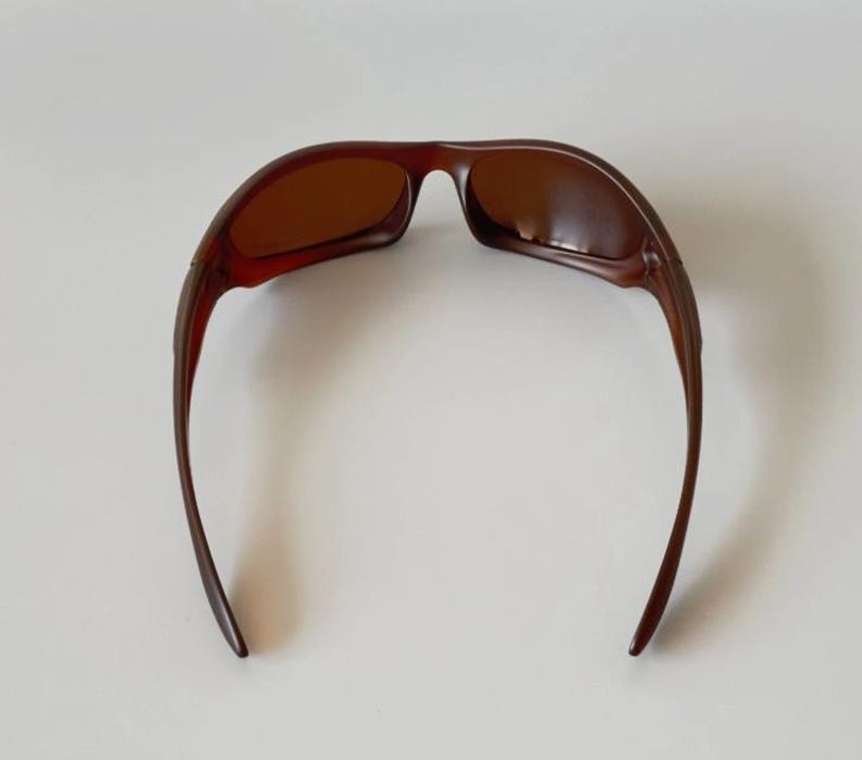 May include: Brown sunglasses with a curved frame and tinted lenses. The sunglasses have a sleek, modern design, with a dark brown color. The arms of the sunglasses curve down and away from the lenses.