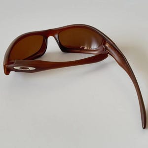 May include: Brown sunglasses with a curved frame and tinted lenses. The sunglasses have a sleek, modern design, with a small silver emblem on the side of the frame. The lenses are a warm brown color, and the frame is a darker shade of brown.
