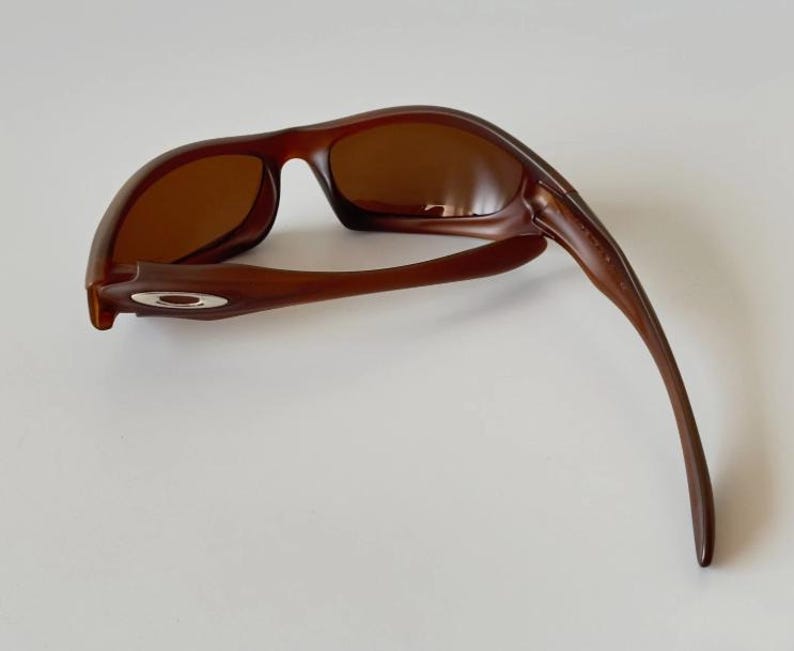 May include: Brown sunglasses with a curved frame and tinted lenses. The sunglasses have a sleek, modern design, with a small silver emblem on the side of the frame. The lenses are a warm brown color, and the frame is a darker shade of brown.