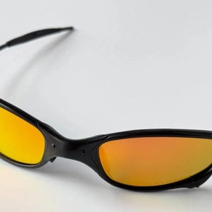 May include: Black sunglasses with orange-yellow lenses. The sunglasses have a sleek, sporty design with a black frame and curved temples. The lenses have a gradient effect, transitioning from yellow to orange.
