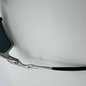 Puede incluir: Primer plano de gafas de sol con montura de metal plateado y lentes azul oscuro. Las patillas son plateadas con una empu&ntilde;adura de goma negra. Sobre un fondo blanco.