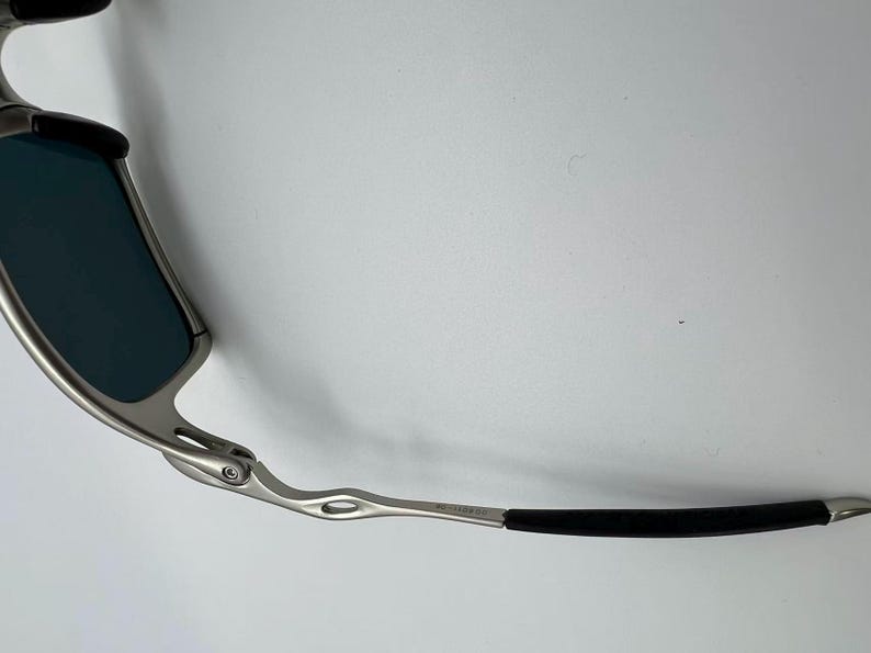 Puede incluir: Primer plano de gafas de sol con montura de metal plateado y lentes azul oscuro. Las patillas son plateadas con una empu&ntilde;adura de goma negra. Sobre un fondo blanco.