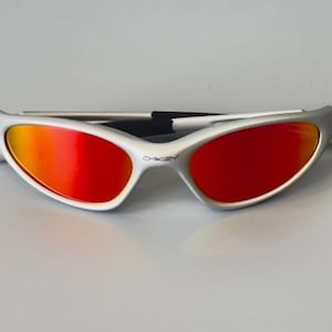 K&ouml;nnte beinhalten: Wei&szlig;-silberne Sonnenbrille mit leuchtend roten und orangefarbenen Gl&auml;sern. Die Sonnenbrille hat ein schlankes, sportliches Design mit einem geschwungenen Rahmen und einem schwarzen Steg. Das Oakley-Logo ist sichtbar.