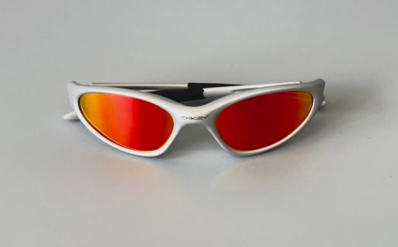 K&ouml;nnte beinhalten: Wei&szlig;-silberne Sonnenbrille mit leuchtend roten und orangefarbenen Gl&auml;sern. Die Sonnenbrille hat ein schlankes, sportliches Design mit einem geschwungenen Rahmen und einem schwarzen Steg. Das Oakley-Logo ist sichtbar.