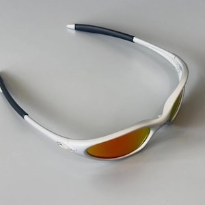 K&ouml;nnte beinhalten: Wei&szlig; gerahmte Sonnenbrille mit orangefarbenen Gl&auml;sern. Die B&uuml;gel sind wei&szlig; mit schwarzen Gummispitzen. Die Gl&auml;ser haben eine verspiegelte Oberfl&auml;che. Geeignet f&uuml;r Sport und Outdoor-Aktivit&auml;ten.