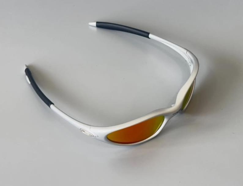 K&ouml;nnte beinhalten: Wei&szlig; gerahmte Sonnenbrille mit orangefarbenen Gl&auml;sern. Die B&uuml;gel sind wei&szlig; mit schwarzen Gummispitzen. Die Gl&auml;ser haben eine verspiegelte Oberfl&auml;che. Geeignet f&uuml;r Sport und Outdoor-Aktivit&auml;ten.