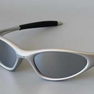 Puede incluir: Gafas de sol plateadas con lentes oscuras. Las gafas de sol tienen un diseño elegante y moderno con monturas curvas y detalles negros en las patillas. El logotipo de Oakley es visible en la montura.
