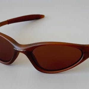 Puede incluir: Gafas de sol marrones con montura curvada y lentes tintadas. Las gafas de sol tienen un diseño elegante y deportivo, con el nombre de la marca visible en la montura. Las patillas son de color marrón oscuro.
