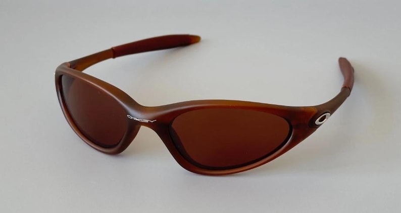 May include: Brown sunglasses with a curved frame and tinted lenses. The sunglasses have a sleek, sporty design, with the brand name visible on the frame. The arms of the sunglasses are a darker shade of brown.