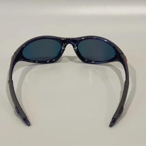 May include: Dark blue sunglasses with a curved frame and dark lenses. The sunglasses have a sleek, sporty design, suitable for outdoor activities. The temples are a darker shade of blue.