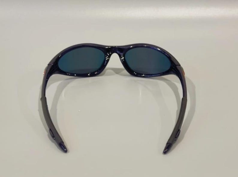 May include: Dark blue sunglasses with a curved frame and dark lenses. The sunglasses have a sleek, sporty design, suitable for outdoor activities. The temples are a darker shade of blue.