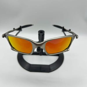 Puede incluir: Gafas de sol con montura plateada y lentes naranjas. Se exhiben sobre un soporte negro sobre un fondo blanco. Las lentes presentan un degradado de color, pasando de un naranja brillante a un tono m&aacute;s oscuro.