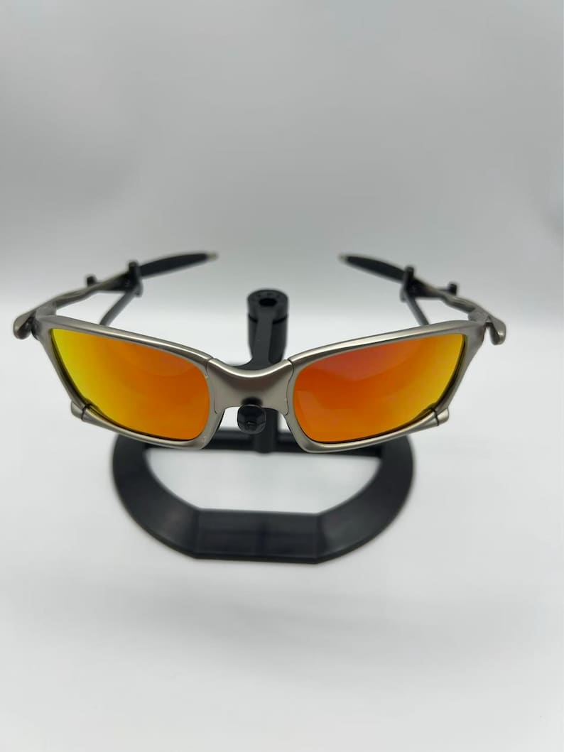 Puede incluir: Gafas de sol con montura plateada y lentes naranjas. Se exhiben sobre un soporte negro sobre un fondo blanco. Las lentes presentan un degradado de color, pasando de un naranja brillante a un tono m&aacute;s oscuro.