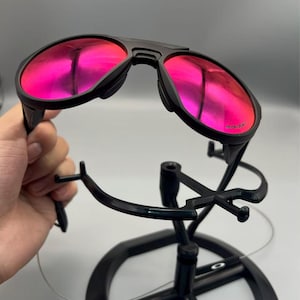 May include: Black sunglasses with pink mirrored lenses. The sunglasses have a double bridge design and are held by a black display stand. The lenses reflect a bright pink colour.