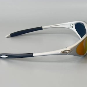 K&ouml;nnte beinhalten: Silberne Sonnenbrille mit schwarzen B&uuml;geln und blau-orangen Gl&auml;sern. Die Sonnenbrille hat ein sportliches Design. Das Oakley-Logo ist auf dem Rahmen sichtbar. Geeignet f&uuml;r Outdoor-Aktivit&auml;ten.